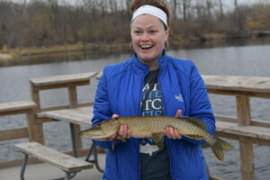 Lauren Jescovitch smiles and holds a northern pike in her hands while standing on a fishing dock in Bay City State Park