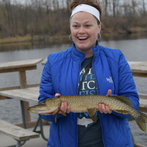 Lauren Jescovitch smiles and holds a northern pike in her hands while standing on a fishing dock in Bay City State Park