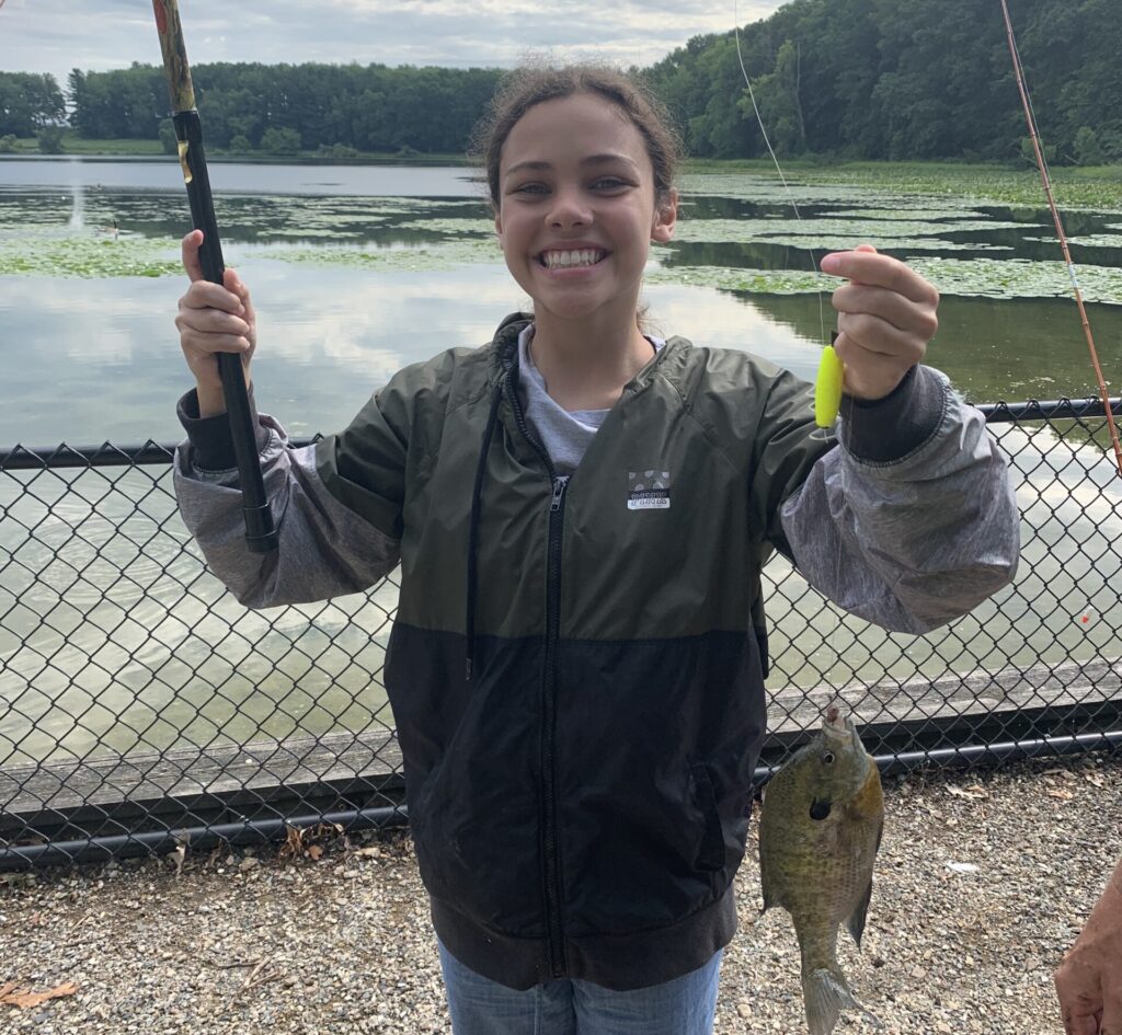 A camper proudly smiles with their freshly caught fish next to a pond