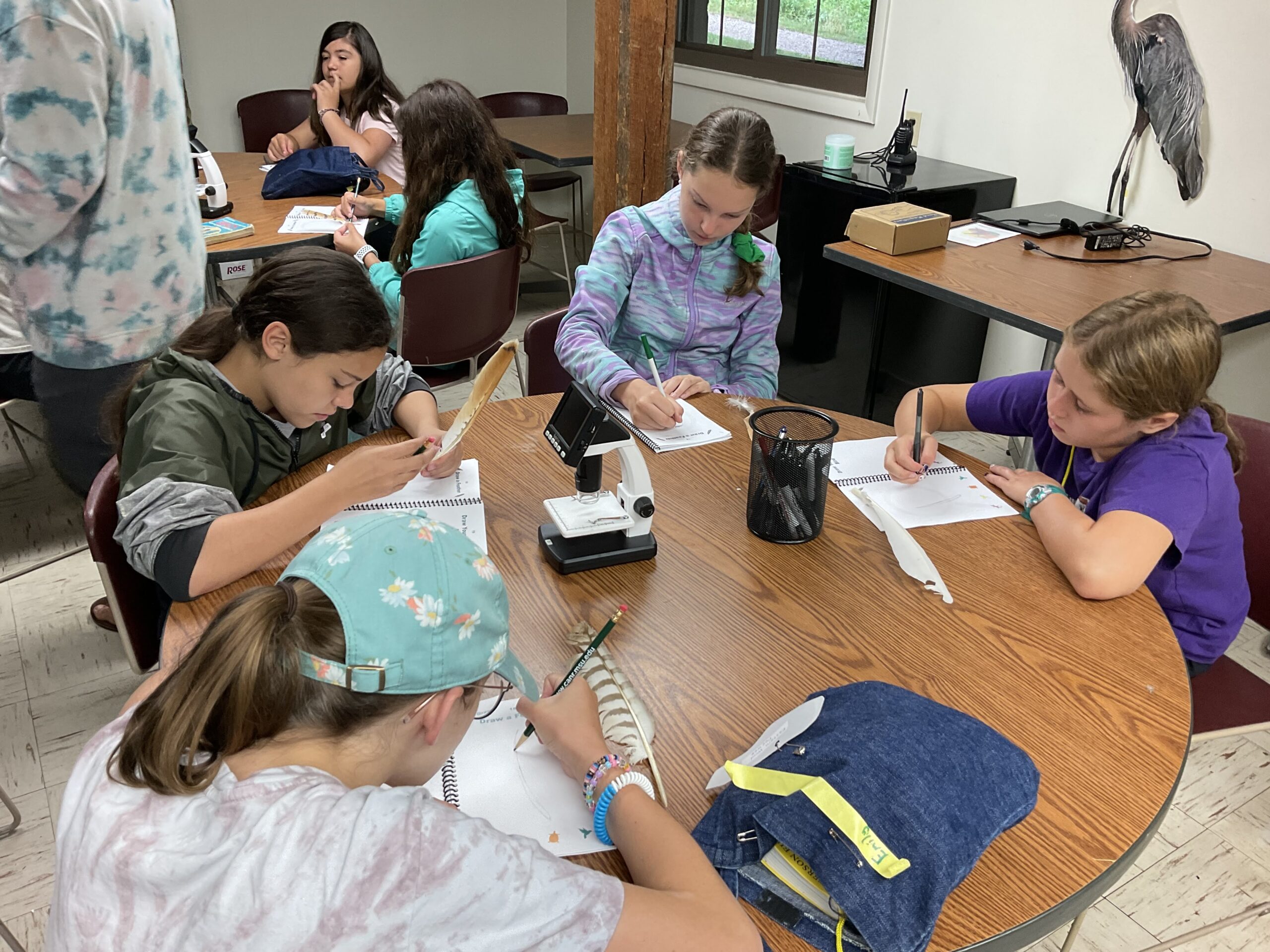 A group of campers sit at a table holding feathers and writing in notebooks