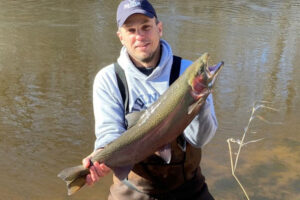 Man wearing waders is seen standing in the river holding a steelhead he caught.