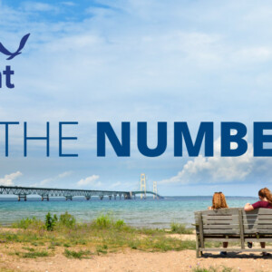 By the numbers graphic with two people sitting on a bench looking at the mackinac bridge on Lake Huron