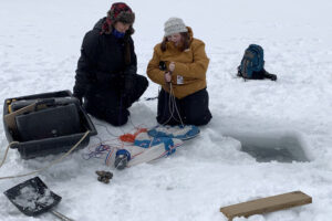 Two researchers sit on an ice covered lake with a hole to the right, and research equipment nearby