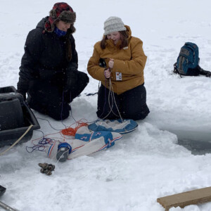 Two researchers sit on an ice covered lake with a hole to the right, and research equipment nearby