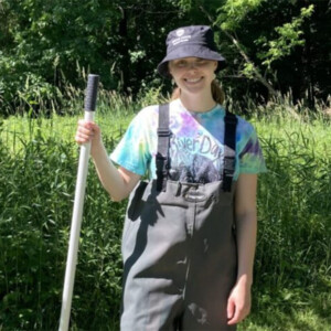 Ella stands in a wooded area, wearing waders and holding a dip net.