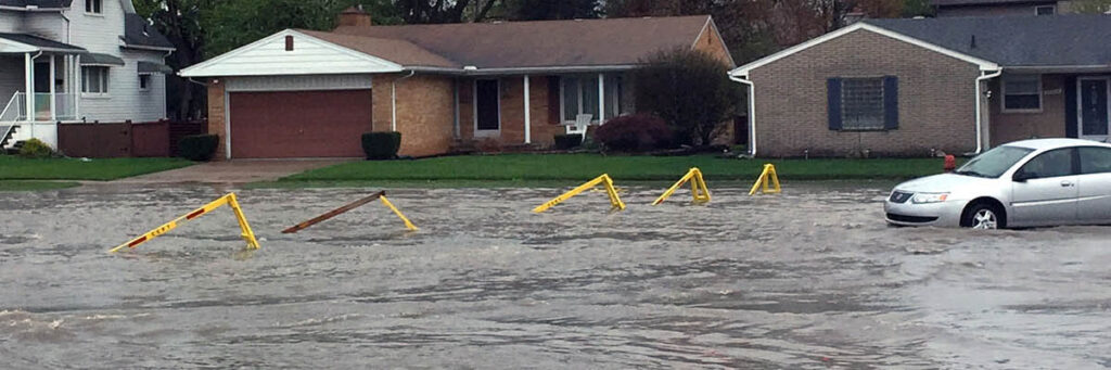 A car drives into flooded street with homes in the background