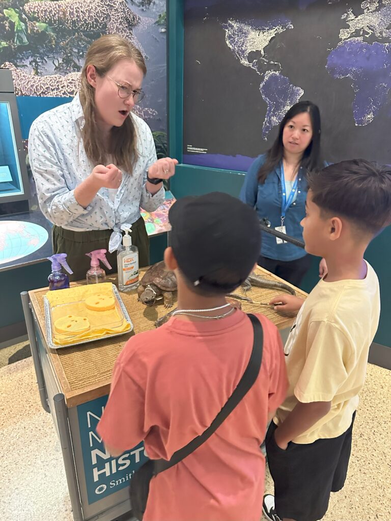 Brenna, a blonde white woman, speaks with two children about amphibians at a display booth