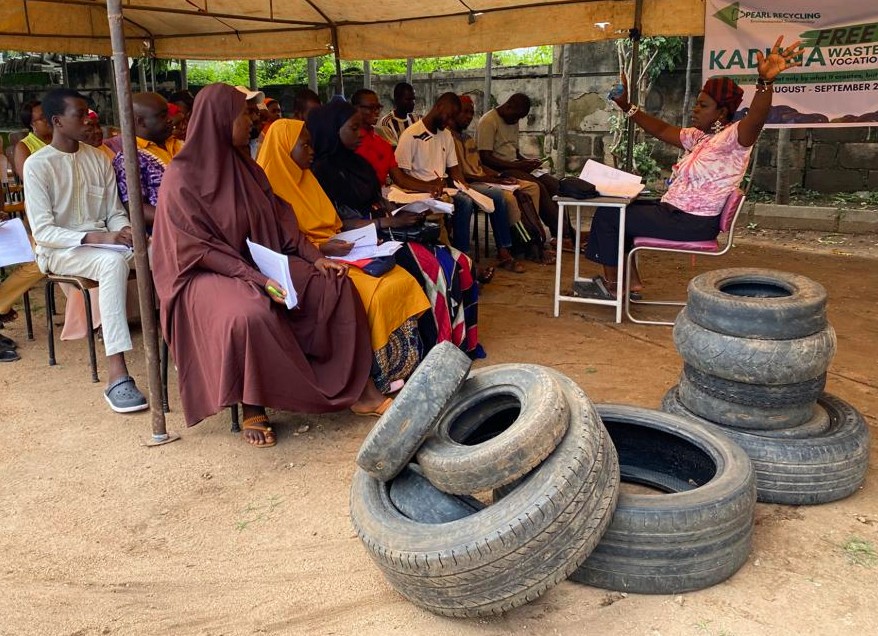 A group of Nigerian people sit under a tent at a training workshop beside a small pile of rubber tires.