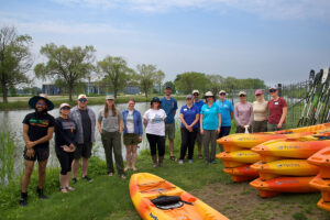 A group of 15 Mi Paddle Stewards stand near a canal on Belle Isle with Kayaks in the foreground.