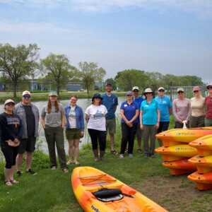 A group of 15 Mi Paddle Stewards stand near a canal on Belle Isle with Kayaks in the foreground.