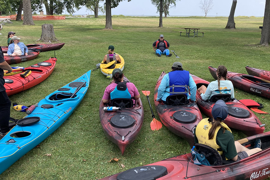 Kayak safety training session with people sitting in kayaks in the grass learning maneuvers with the Lake in the background
