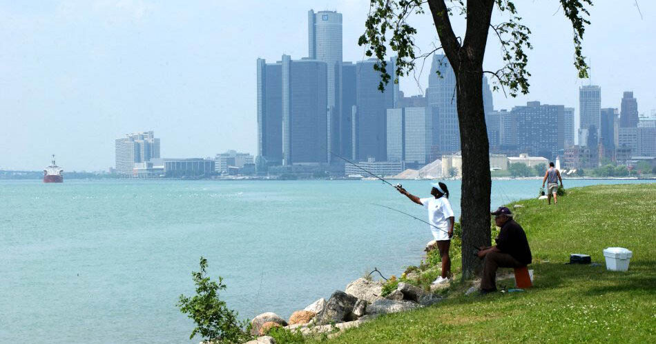 2 people fishing along a wide river with a city, bridge, and freighter in the background