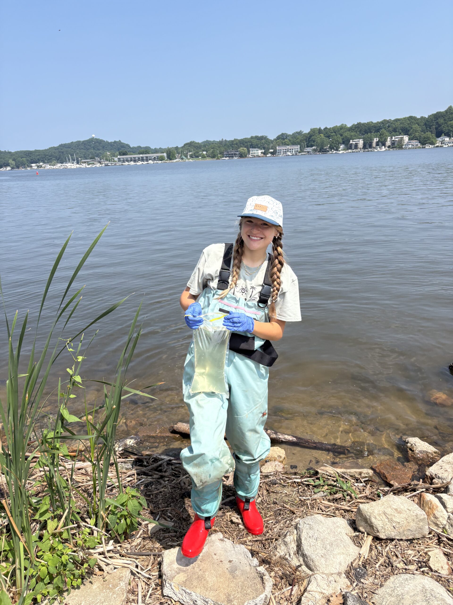 Annie stands in shallow waters at the edge of a lake, holding a sample bag of water