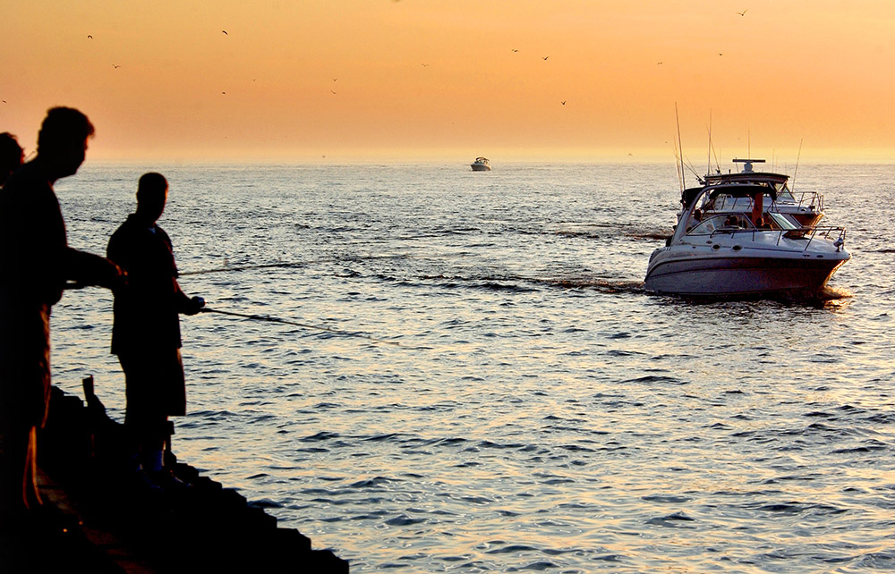 fishers on a pier with a recreation boat passing by in a channel.