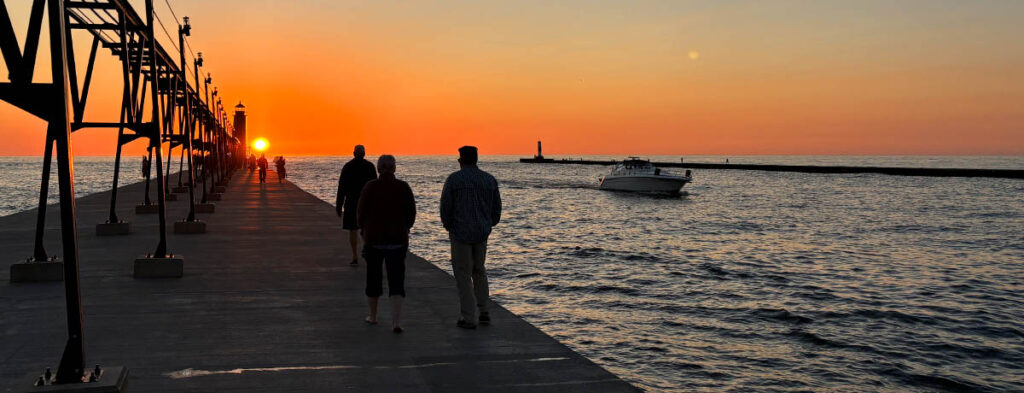 sunset from a pier with people walking along the boardwalk, and a boat in the channel