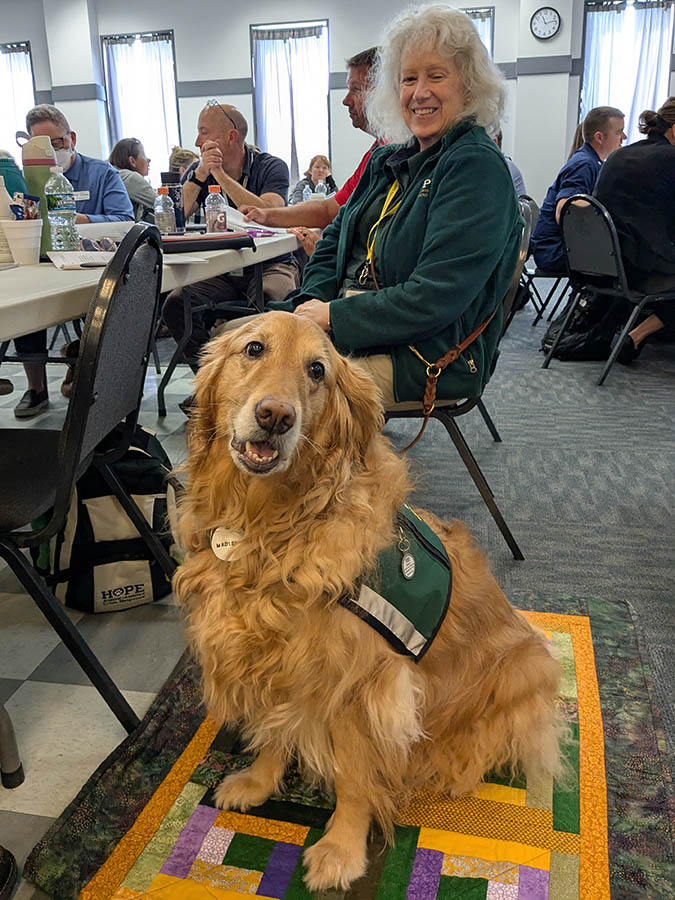 A therapy dog sits on a quilt near her human handler during a meeting