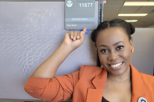 Ola Ayeni, a Black woman in an orange blazer, smiles as she points at an office nameplate bearing her name