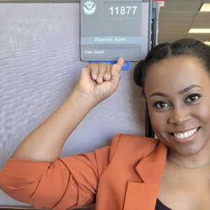 Ola Ayeni, a Black woman in an orange blazer, smiles as she points at an office nameplate bearing her name