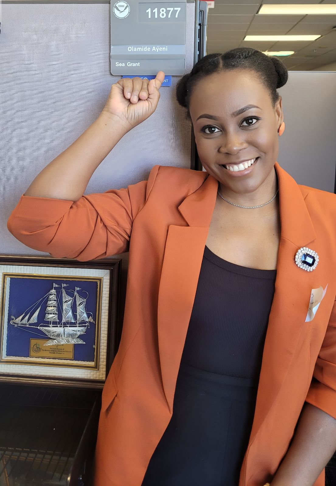 Ola Ayeni, a Black woman in an orange blazer, smiles as she points at an office nameplate bearing her name