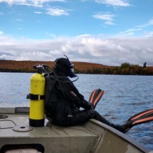 a scuba diver in a black suit with a yellow oxygen tank looks out onto the lake on a colorful fall day