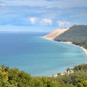 A large dune that slopes down do a blue-green body of water with a partly sunny sky, a forest is seen in the foreground