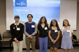 A group of five students pose for a photo during a symposium in front of a screen that has a photo of sand dunes along a lake
