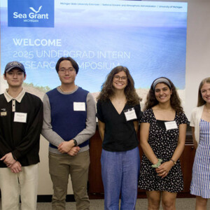 A group of five students pose for a photo during a symposium in front of a screen that has a photo of sand dunes along a lake