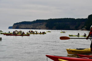 A very large group of kayaks on Lake Superior with Pictured Rocks National Lakeshore in the background on a cloudy day