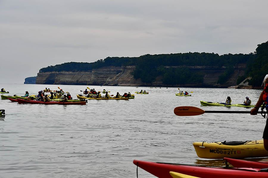 A very large group of kayaks on Lake Superior with Pictured Rocks National Lakeshore in the background, on a cloudy day