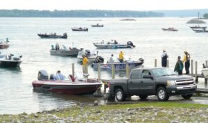 A number of fishing boats are seen on a river. A pickup truck is backed up to the lake at a boat launch and launching a boat .