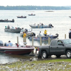 A number of fishing boats are seen on a river. A pickup truck is backed up to the lake at a boat launch and launching a boat .