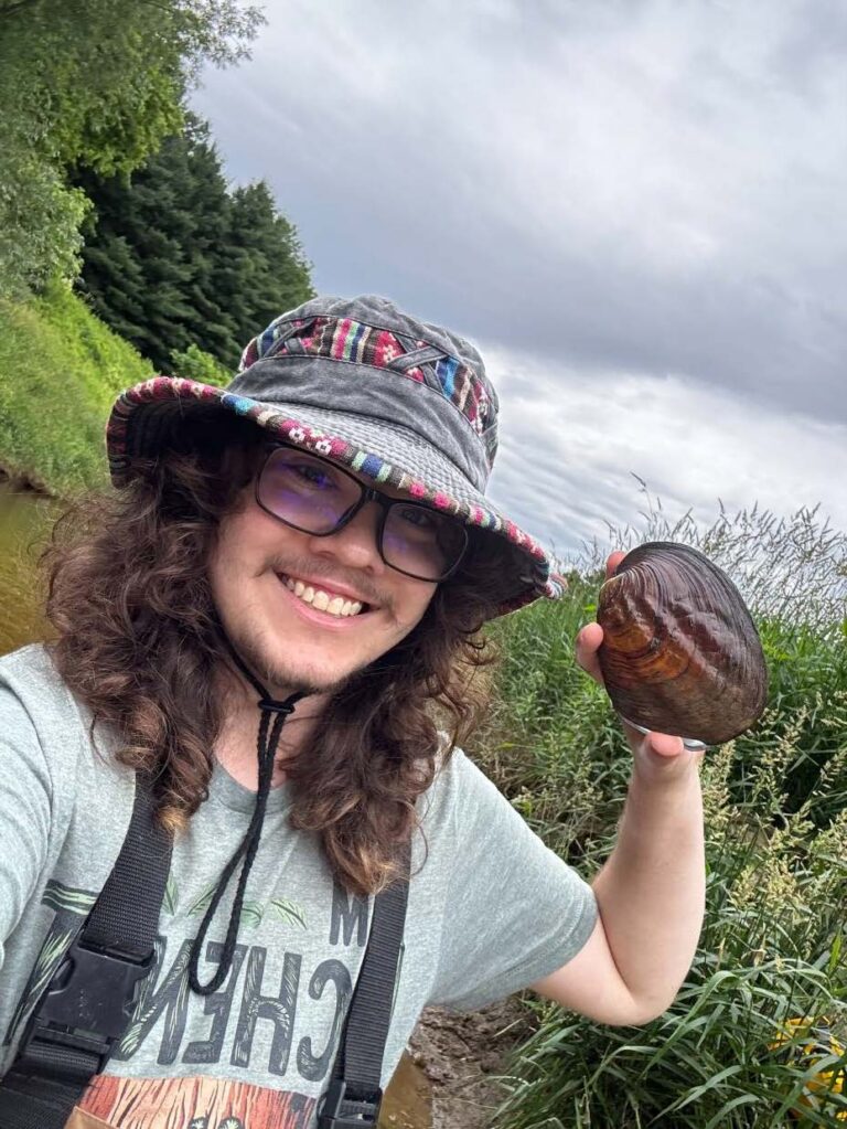 A young researcher smiles while holding up a large native mussel shell