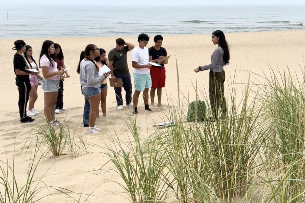 A group of students listen to a instructor on a beach near dunegrass, with a lake in the background.