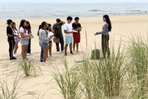 A group of students listen to a instructor on a beach near dunegrass, with a lake in the background.
